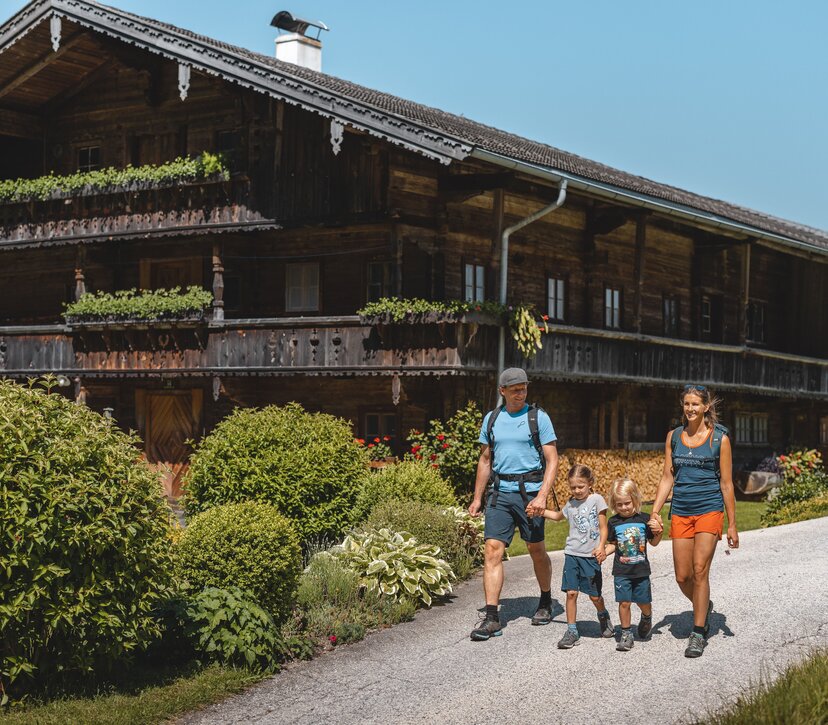 Eine Familie wandert der Straße entlang, vorbei an einem schönen alten Bauernhaus, welches umgeben ist von blühender Natur  | © Alpbachtal Tourismus | Mathäus Gartner 