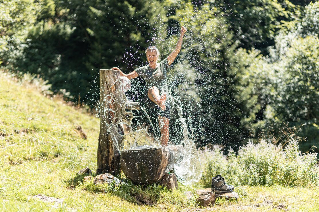 Eine Frau steigt mit dem Fuß in den Brunnen vor der Holzalm und spritzt mit dem Wasser und lacht | © Alpbachtal Tourismus | shootandstyle