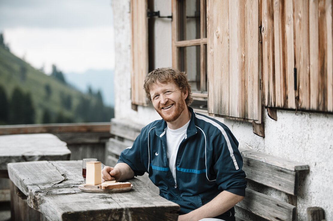 Auf diesem Bild zu sehen ist der Käsemeister Hannes Klingler auf seiner Steinbergalm in Inneralpbach, vor ihm ein Holzbrett mit Käse und Brot sowie ein Glas Rotwein | © Alpbachtal Tourismus | shootandstyle 